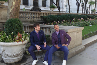 Two men in suits sitting on a bench in front of a building with trees and plants.