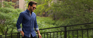 A man wearing a navy blue long-sleeve button-down shirt with rolled-up cuffs and dark sunglasses, walking outdoors on a balcony with a green, tree-filled background.