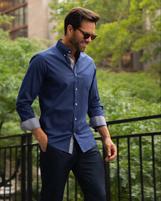 A man wearing a blue gingham long-sleeve button down shirt with light blue rolled cuffs, paired with dark dress pants, standing outdoors on a walkway with green foliage in the background.