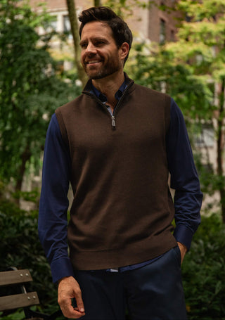 A man wearing a brown quarter-zip knit sweater vest over a navy blue long-sleeve shirt, standing outdoors in a park-like setting with trees and a bench in the background.