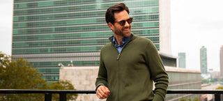 A man wearing a green quarter-zip sweater over a blue collared shirt, standing outdoors on a balcony with a modern glass building in the background.