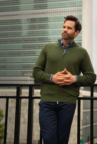 A man wearing an olive green zip-up sweater over a blue collared shirt and navy pants, standing outdoors in front of a modern glass building.
