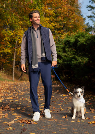 A man wearing a navy blue vest, performance quarter zip, and navy performance pants walks a white and gray dog on a blue leash along an autumn tree-lined path.
