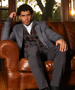 A man wearing a gray suit and patterned vest sits on a brown leather couch in a sunlit room with large windows and potted plants.