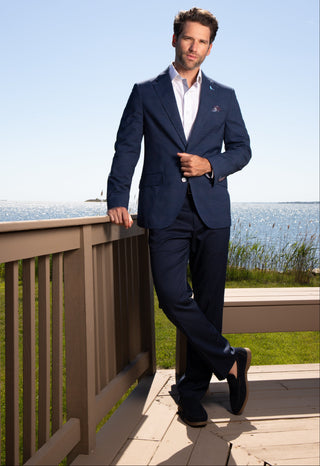 A man wearing a navy blue suit, white dress shirt, and a small red pocket square stands on a wooden deck with a lake and grassy shoreline in the background.