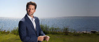 A man in a navy blue suit, white dress shirt, and patterned tie stands outdoors on a grassy area with water and reeds in the background.
