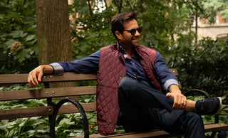 A man wearing a burgundy quilted vest over a blue shirt and dark pants sits on a wooden park bench surrounded by green trees and foliage. 