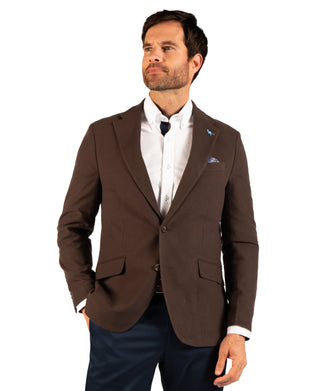 A man wearing a brown tailored blazer over a white dress shirt and dark tie, paired with navy trousers, photographed against a plain white studio background.