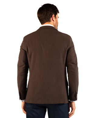 A man viewed from the back wearing a dark brown blazer over a white collared shirt and navy trousers, isolated on a white background.