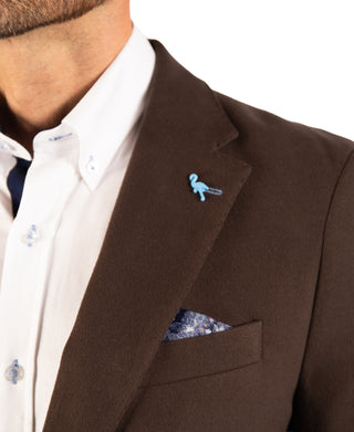 A close-up of a man wearing a brown wool-blend suit jacket over a white button-down shirt. The jacket features a notch lapel decorated with a small light blue bird pin, and a matching blue and white patterned pocket square is tucked into the breast pocket.