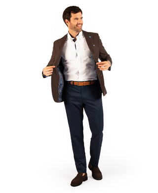 A man modeling a men's suit consisting of a brown blazer with a blue paisley lining, a white dress shirt, navy blue trousers, a brown belt, and brown suede loafers, against a solid white background.