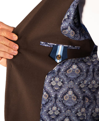 Close-up of a brown tailored jacket or vest being held open by a hand, showing a patterned blue and beige lining with a decorative buttoned pocket flap.