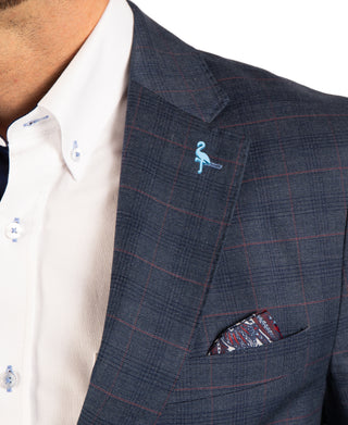 Close-up of a man wearing a navy blue windowpane plaid suit jacket over a white dress shirt, featuring a light blue flamingo lapel pin and a patterned pocket square.