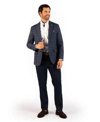 A man modeling a navy blue plaid suit jacket and matching trousers, styled with a white dress shirt, black tie, and brown loafers, against a plain white background.