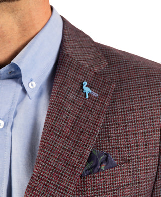 Close-up of a man wearing a brown and red micro-check patterned blazer over a light blue dress shirt, featuring a small light blue flamingo lapel pin and a patterned pocket square.