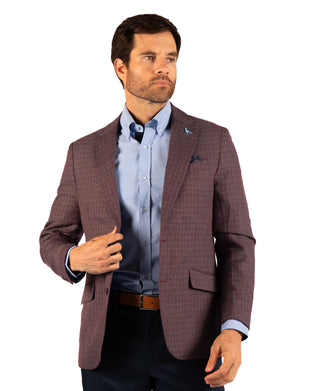 A man wearing a brown and burgundy checkered blazer over a light blue dress shirt, paired with dark trousers and a brown belt, photographed against a solid white background.