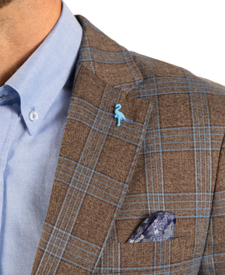 Close-up of a man's neck and chest wearing a brown plaid blazer with a light blue dress shirt, a small light blue bird lapel pin, and a patterned pocket square.