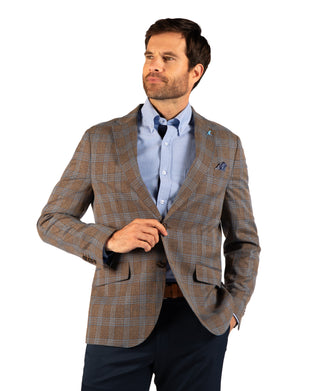 A man wearing a brown and beige plaid blazer over a light blue dress shirt and dark trousers, standing against a plain white background.