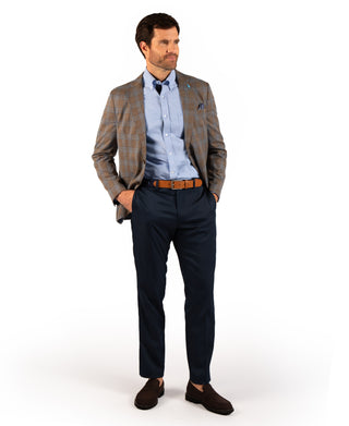 A man modeling a men's outfit consisting of a brown and beige plaid blazer, a light blue dress shirt, a navy blue tie, dark navy trousers, a brown leather belt, and dark brown suede loafers, against a solid white background.