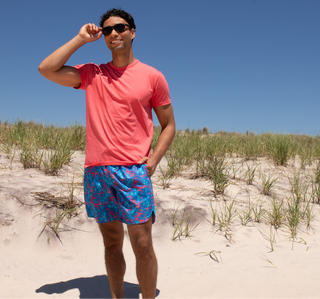 A man wearing a coral pink t-shirt and blue patterned swim trunks stands on a sandy beach with dune grass under a clear blue sky.