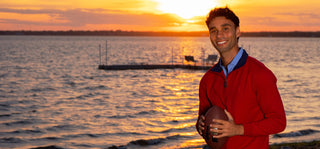 A smiling young man in a red tailorbyrd quarter-zip pullover stands on a lakeshore at sunset, holding a football. The background shows a large body of water with golden sunset reflections and a dock in the distance.