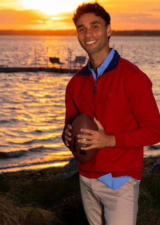A man stands on a grassy shore holding a football, wearing a red quarter-zip sweater over a blue collared shirt, khaki pants, and blue sneakers, with a sunset over water in the background.