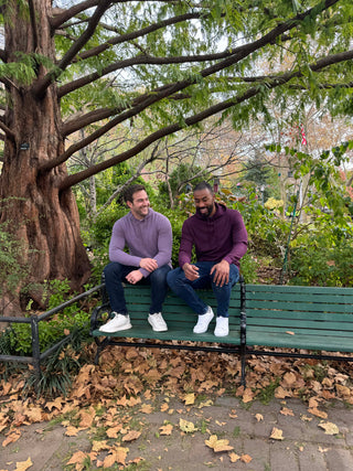 Two men sitting on a bench in a park with trees and fallen leaves.
