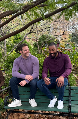 Two men sitting on a bench in a park with trees and greenery in the background.