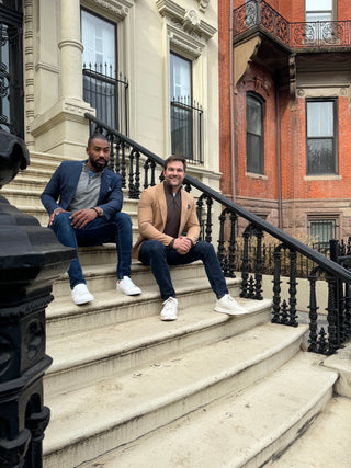 Two men sitting on the steps of a building wearing navy and tan TailorByrd Sport Coats